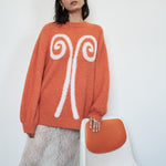 Moody editorial shot of the VARNA Maris Knit Top in tangerine — soft alpaca-wool texture and bold abstract motif.