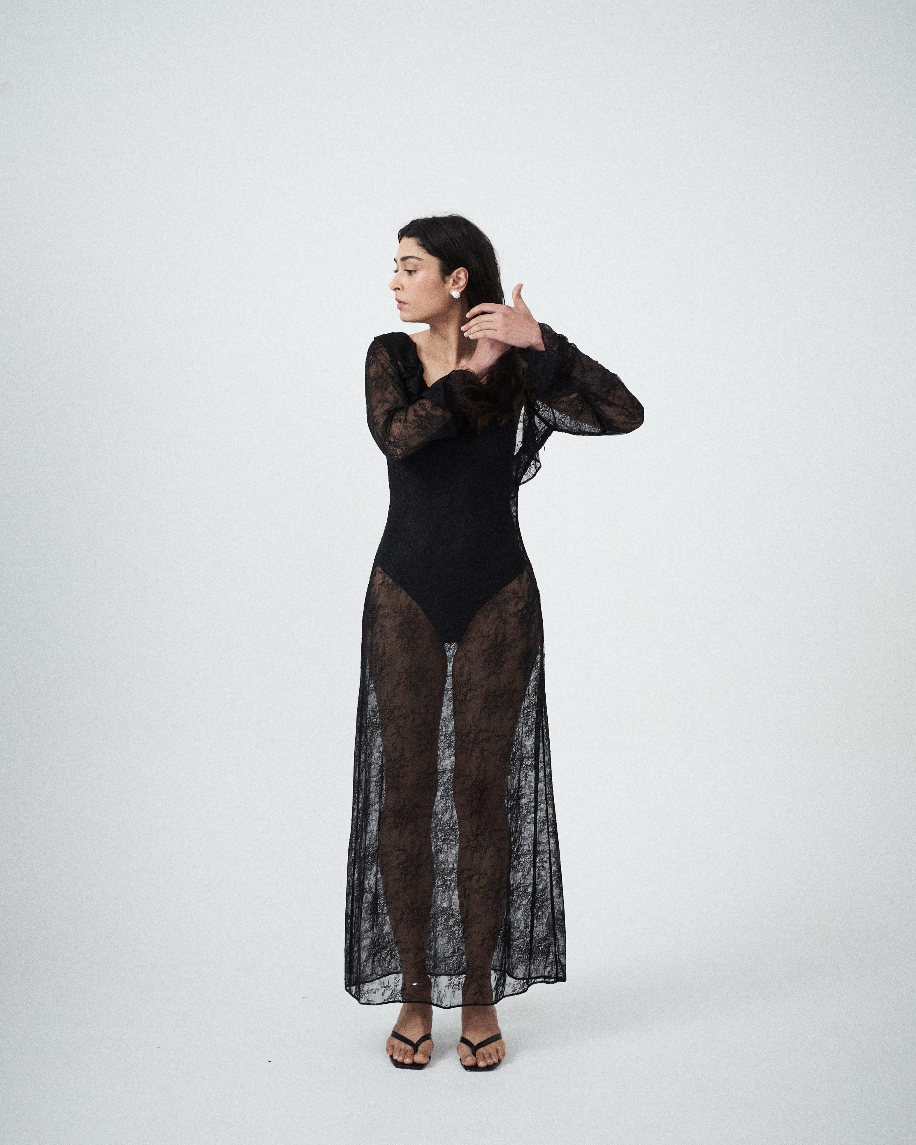 Editorial mood image of the VARNA Lucienne Ruffle Lace Maxi Dress in black — sheer designer lace dress with scoop neckline, long fluted sleeves, and fluid silhouette.