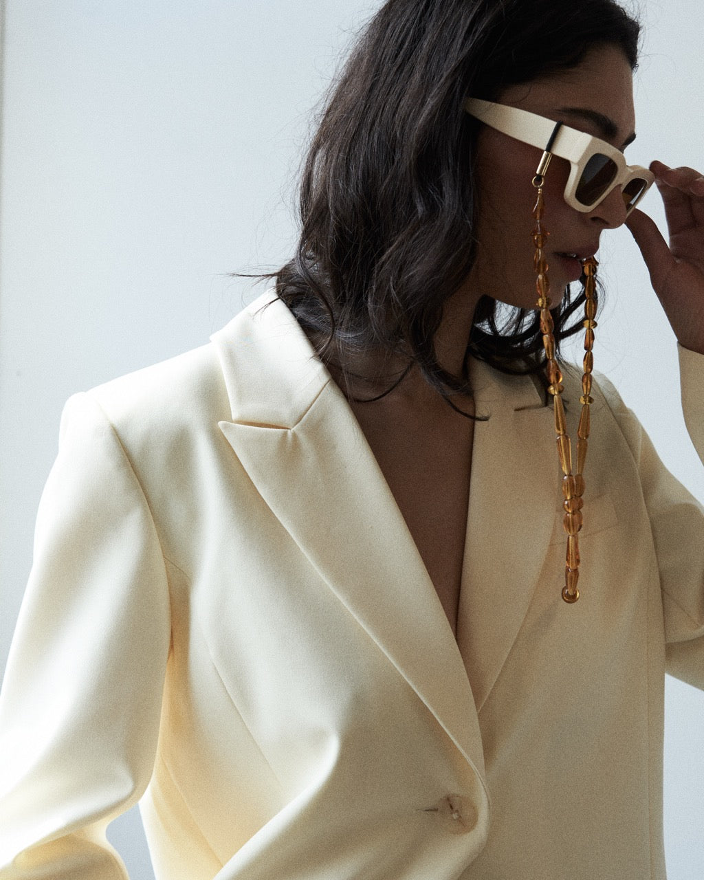 Person wearing a cream blazer and sunglasses with a long chain, against a light background