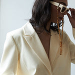 Person wearing a cream blazer and sunglasses with a long chain, against a light background