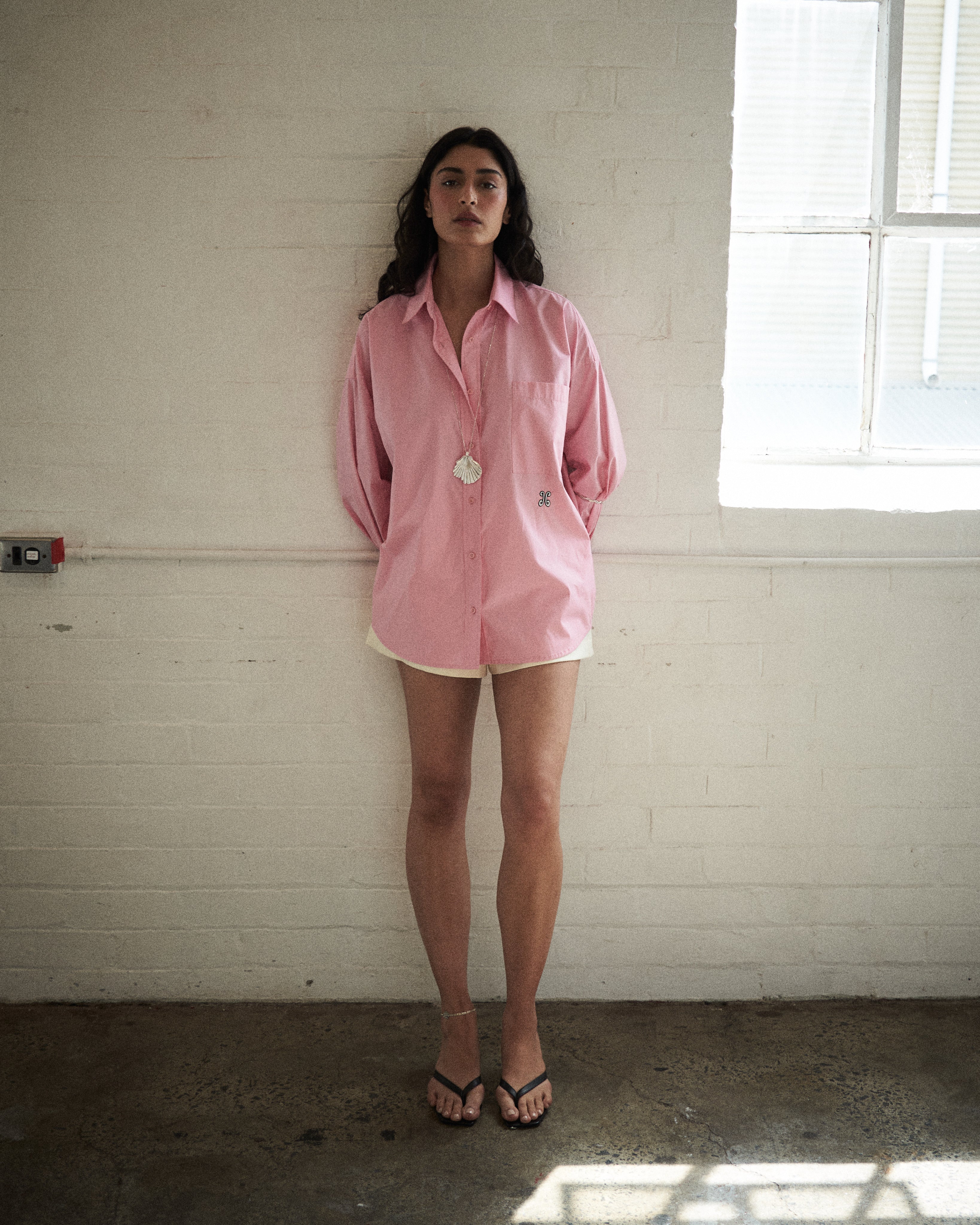 Person wearing a pink shirt, white shorts and a silver shell pendant standing in a room with a window.