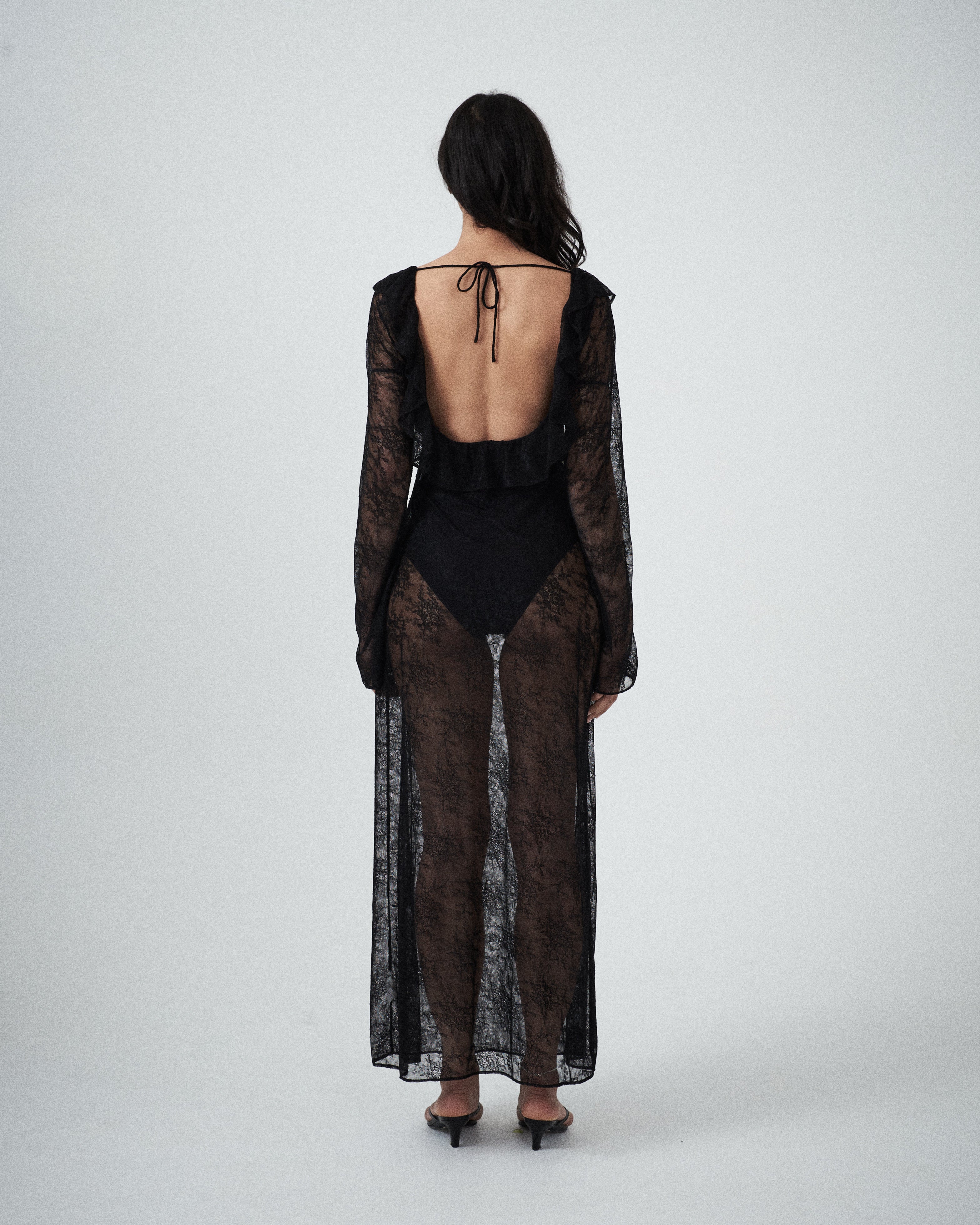 Back view of VARNA Lucienne Ruffle Lace Maxi Dress in black featuring open low back with tie and flowing lace train.